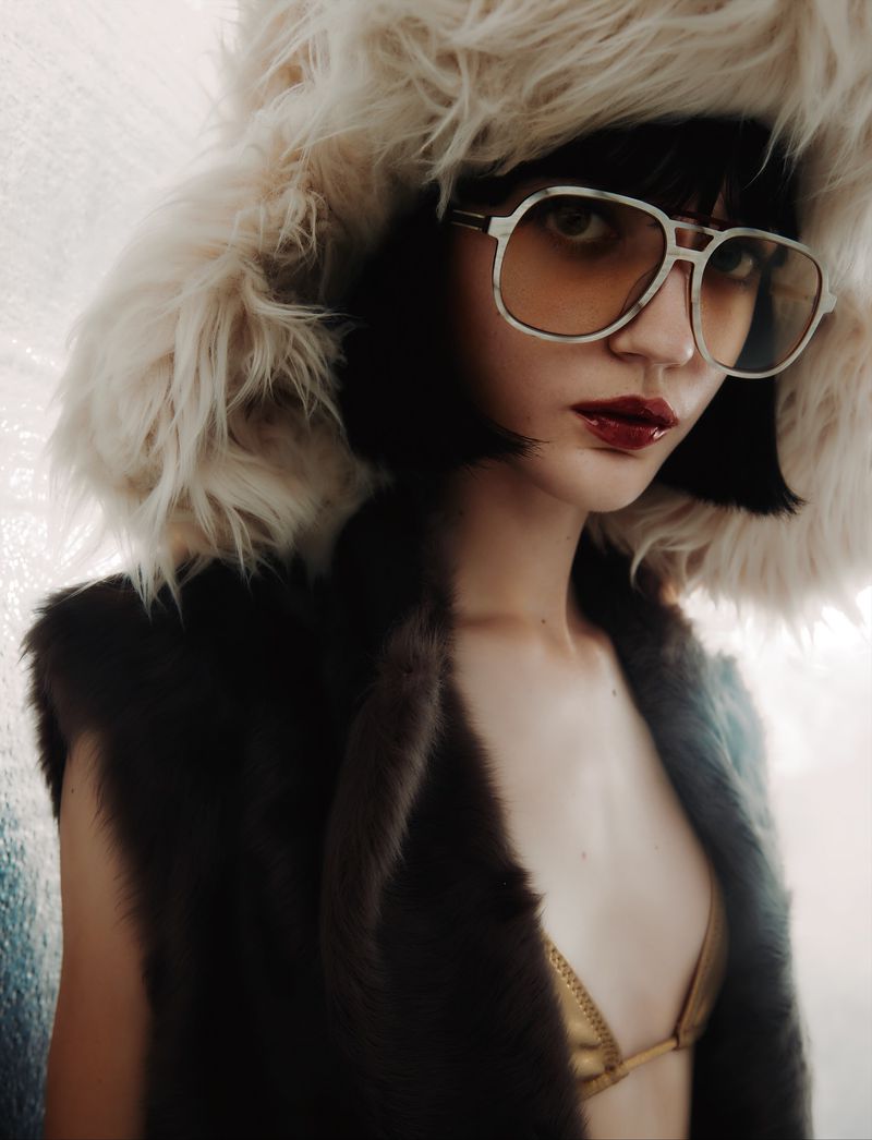 Portrait of a young woman with dark hair wearing a fur hat, a fur vest, a gold bikini top, and retro-style glasses, gazing at the camera.
