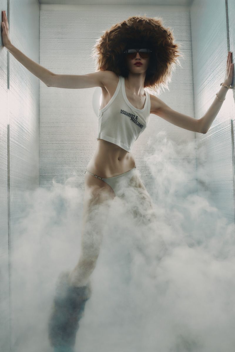 A slender fashion model with a large afro and sunglasses stands in a white tank top amidst smoky haze in a bright studio setting.