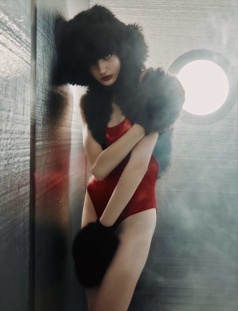 Woman in a red bodysuit poses indoors with a large black fur hat and matching mitts, looking at the camera against a grey wall.