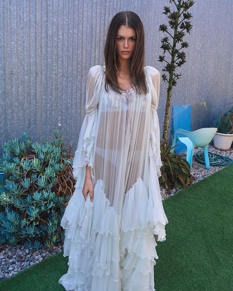 An adult woman stands outdoors wearing a sheer white ruffled dress, with greenery and a corrugated metal fence in the background.