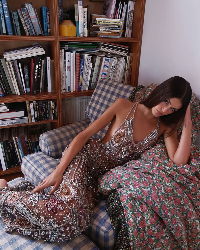 Adult woman in a beaded gown poses on a blue plaid chair with floral pillows in front of a bookshelf inside a house.