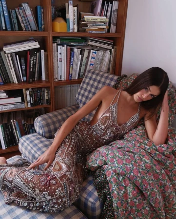 Adult woman in a beaded gown poses on a blue plaid chair with floral pillows in front of a bookshelf inside a house.