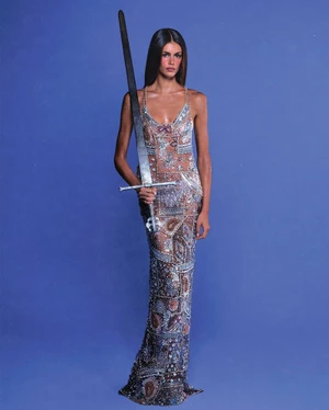Model Kaia Gerber holding a large sword wearing an embellished sheer gown against a blue seamless backdrop for Vanity Fair U.S.