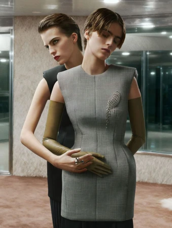 Two women in grey dresses and olive gloves, one with dark lipstick, stand in a studio with metallic accents. Fashion editorial shot.