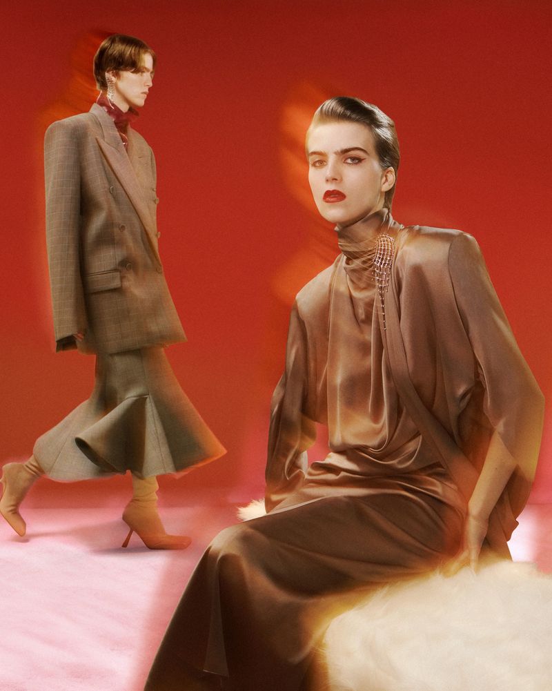 Two women in retro-style high fashion in a studio, one seated in a long dress, the other standing in a suit with a flared skirt.