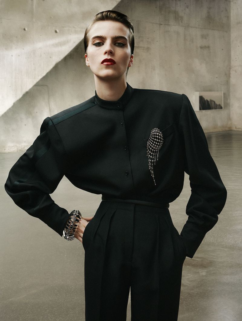 Woman in black tailored shirt and pants with an embellished pin, wearing a cuff bracelet, poses in a minimalist architectural space.