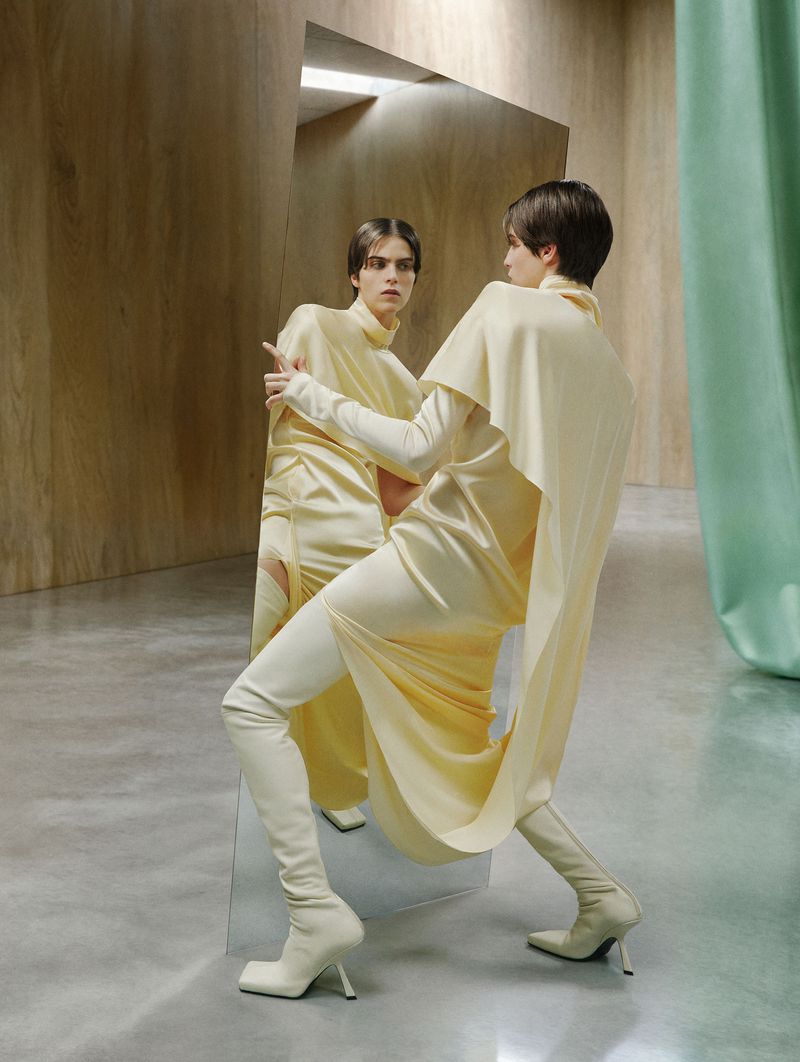 Elegant model poses in a light yellow dress and matching thigh-high boots, reflected in a mirror against a backdrop of minimalist design.
