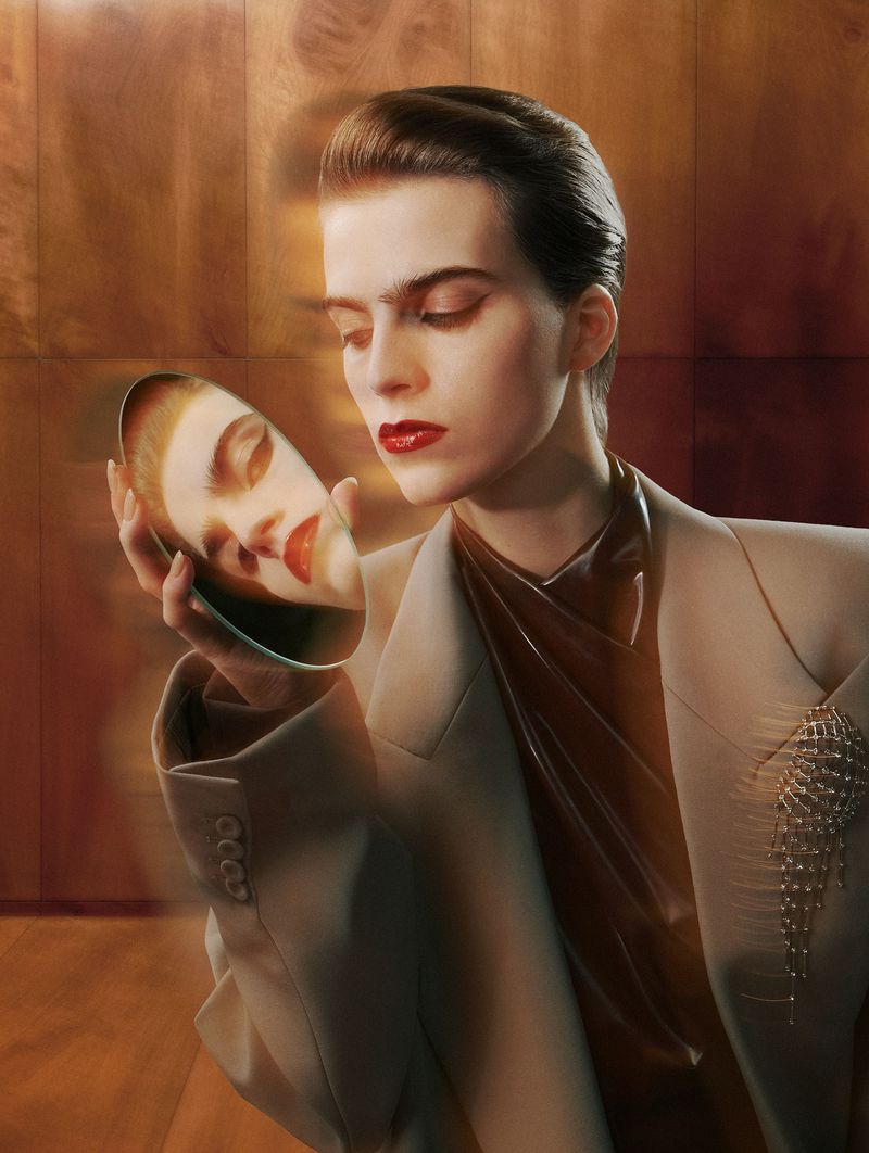 Elegant woman in a cream suit holds a mirror reflecting her face, creating a double portrait against a blurred brown background.