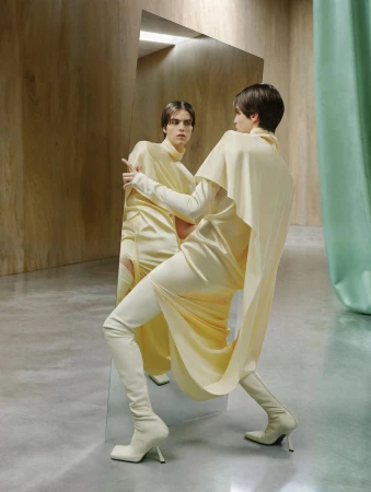 Model in yellow drapery dress and boots posing by a tilted mirror in a wood-paneled room
