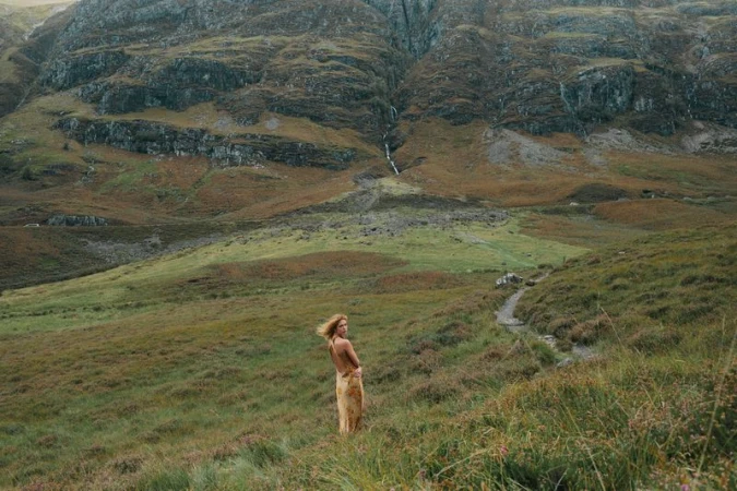 Woman in a tan dress standing in a grassy field with large mountains in the background and a winding path.