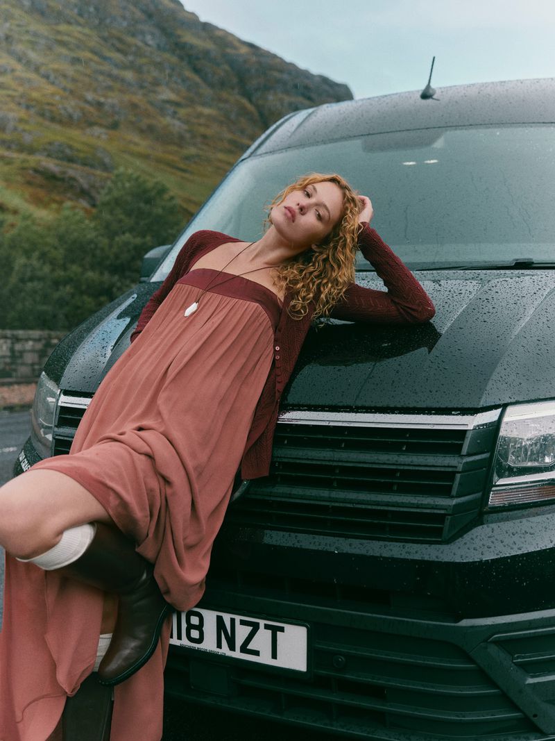 Adult female in maxi dress leans against a black van in an outdoor scene with mountains, wearing boots and a cardigan.