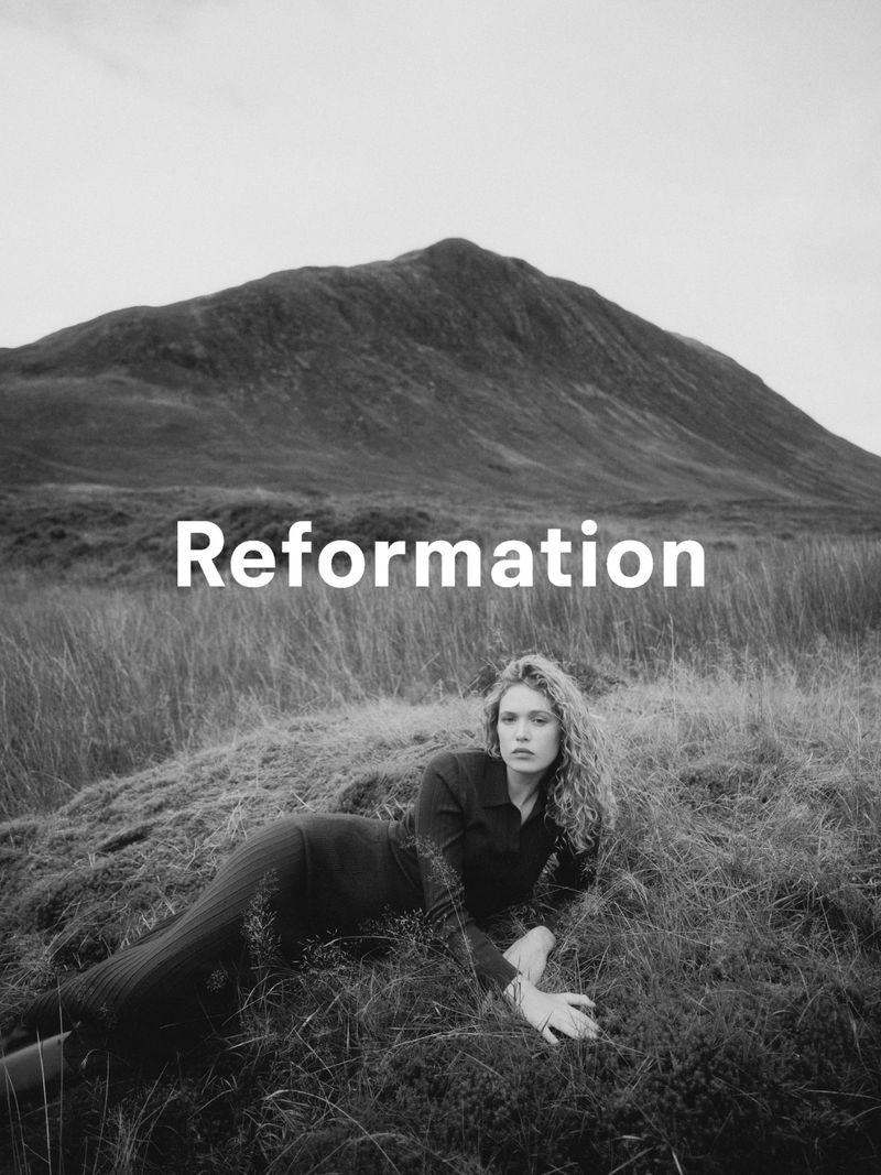 Monochrome fashion shot of a woman lying in a field of grass with a large mountain in the background, fashion brand logo overlayed.