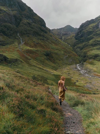 A lone woman in a mustard dress hikes on a rocky path through a lush green valley surrounded by tall, sloping mountains and a cloudy sky.