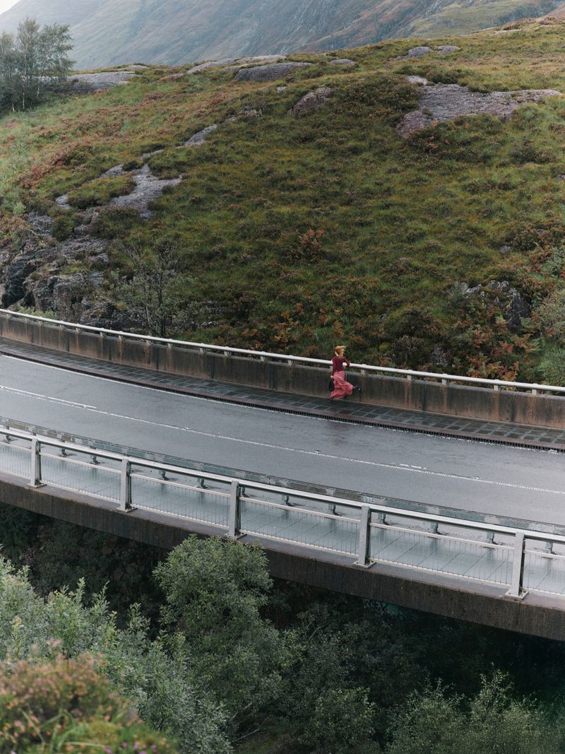 A person walks across a gray concrete bridge in a green, mountainous rural environment on an overcast day, viewed from an elevated angle.