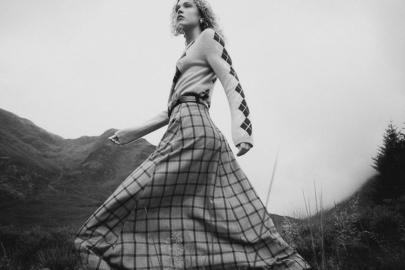 Monochrome fashion shot of a woman with curly hair wearing a patterned sweater and long plaid skirt, walking across a grassy field.