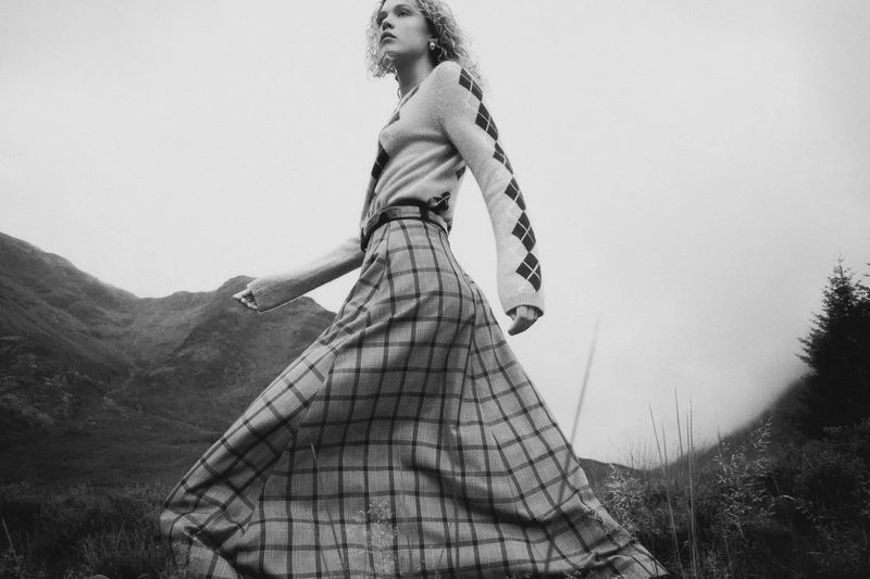 Model wearing patterned argyle sweater and plaid skirt outdoors against mountain landscape background