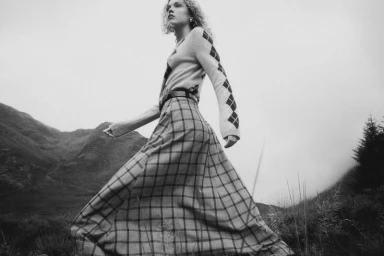 Model wearing patterned argyle sweater and plaid skirt outdoors against mountain landscape background