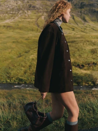 Model with long curly red hair wearing a dark brown jacket and knee-high boots holding a dark handbag in a grassy landscape