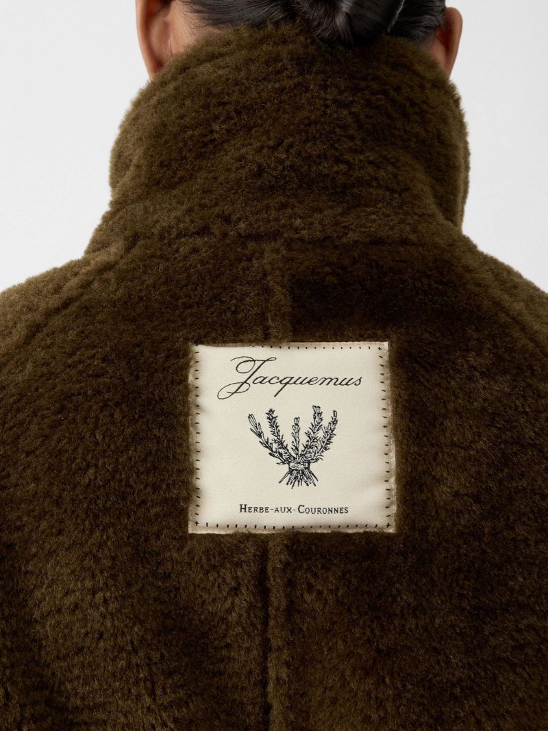 Close-up of a person wearing a brown faux fur jacket, label with Jacquemus logo and plant illustration stitched on the back.