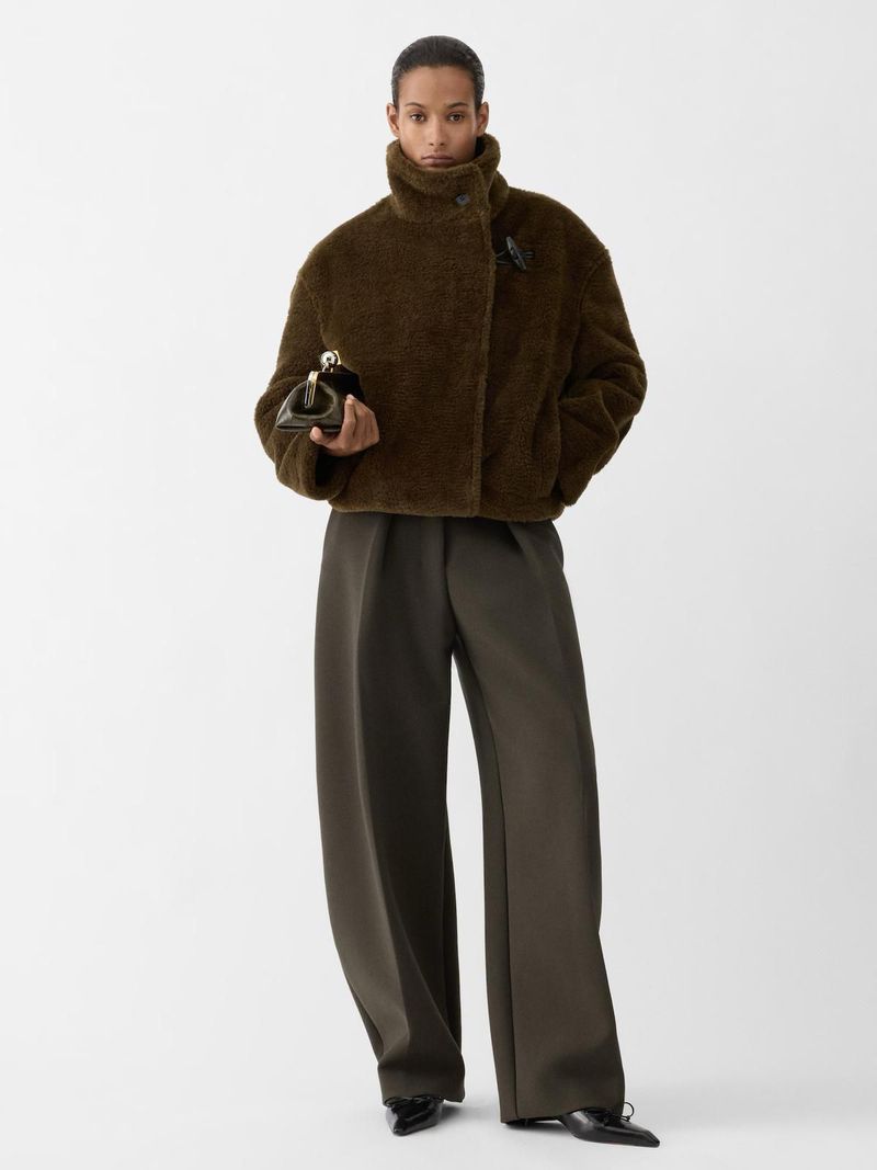 An adult female fashion model poses in a studio wearing a brown jacket and wide-leg trousers, holding a clutch.