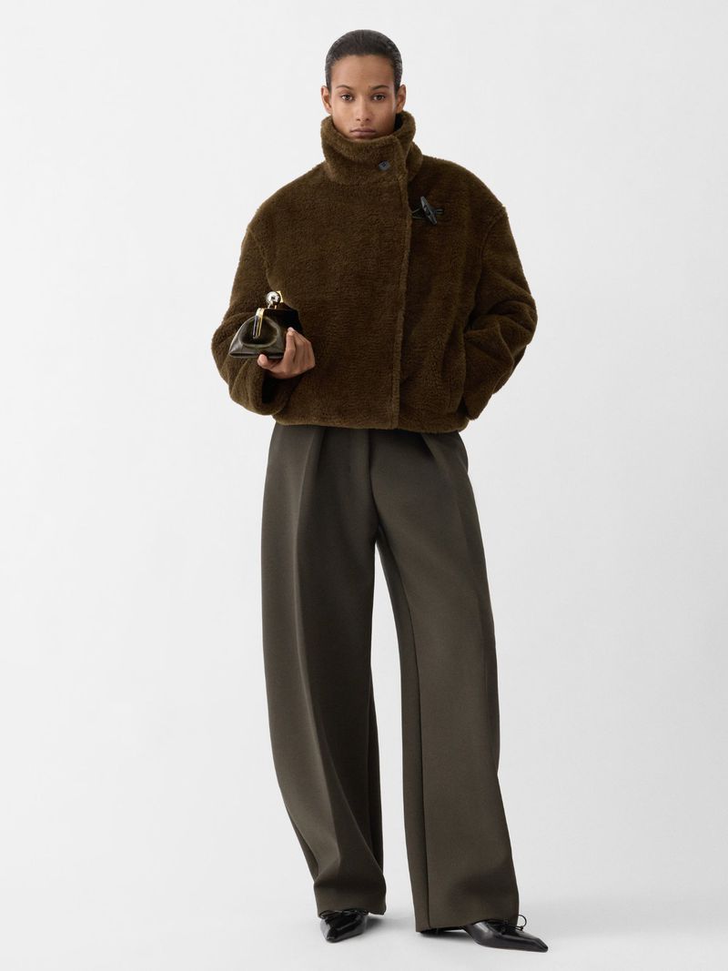 A model poses in a studio wearing an olive-green, teddy-style jacket, matching wide-legged trousers, and black pointed shoes, holding a clutch.