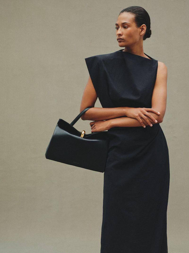 Adult woman in a black dress holds a black leather purse with a gold handle, posing in a neutral-toned studio for a fashion shot.