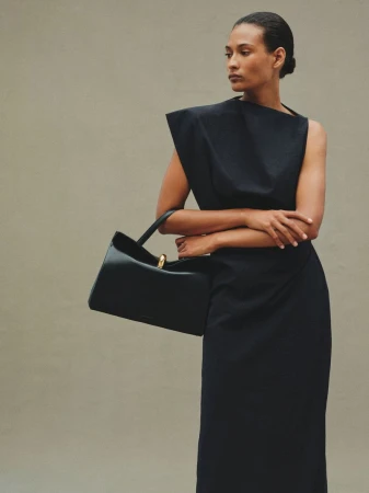 Adult woman in a black dress holds a black leather purse with a gold handle, posing in a neutral-toned studio for a fashion shot.