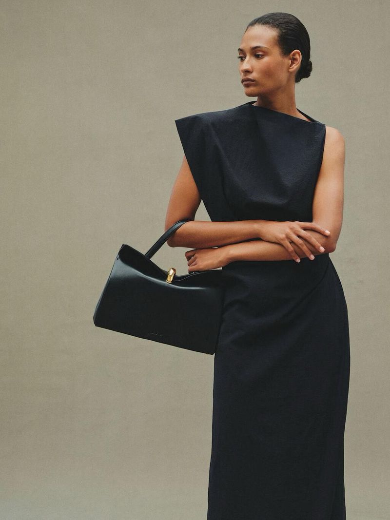 Model wearing a black dress holding a black leather handbag against a neutral grey studio background