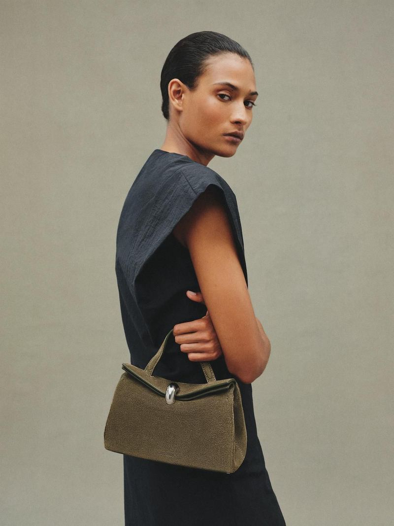 Portrait of a woman in a black dress holding an olive-green handbag with silver hardware against a neutral background in a fashion editorial.