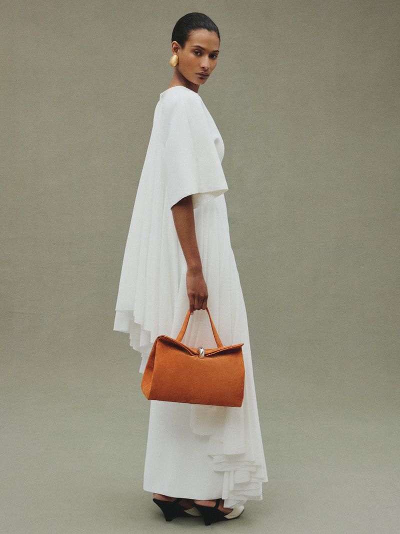 Elegant woman in a white dress with a pleated design and orange handbag, posing in a minimalist studio setting, looking at the camera.