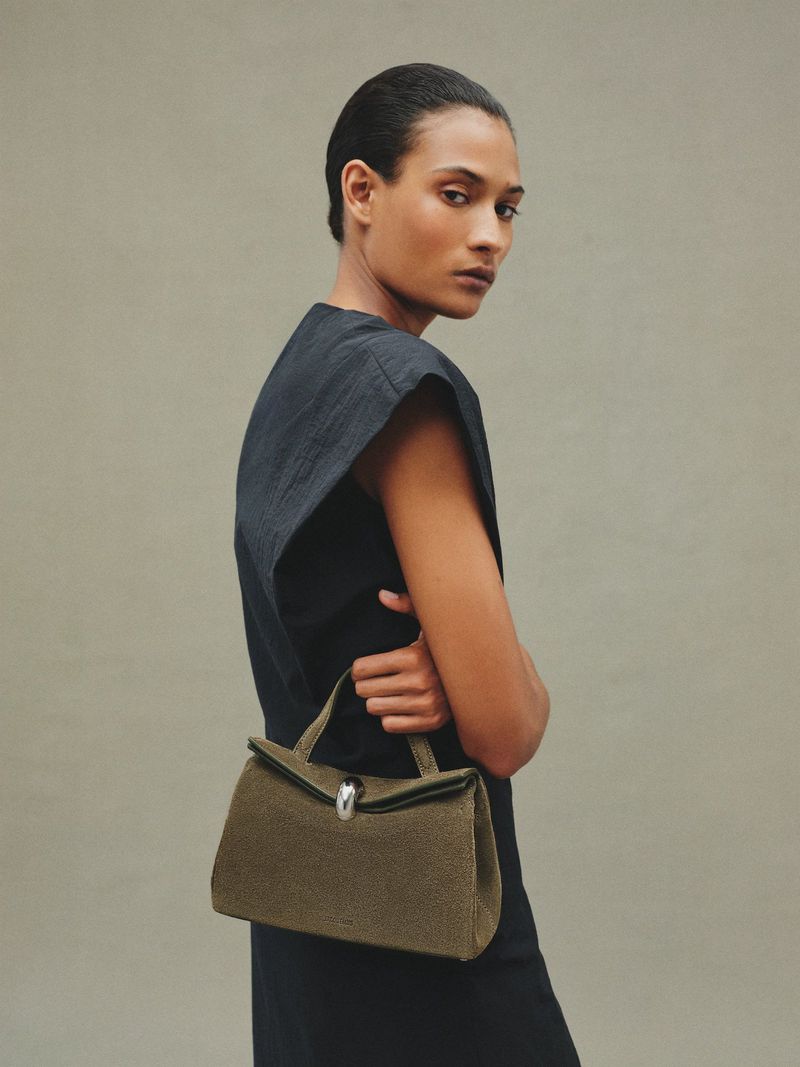 Woman in a black dress holds a green handbag against a neutral background, showcasing fashion and style in a studio setting.