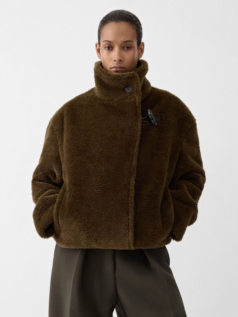 Studio shot of an adult female fashion model wearing a brown, high-necked, sheepskin-textured jacket and olive-green wide-leg trousers on white.