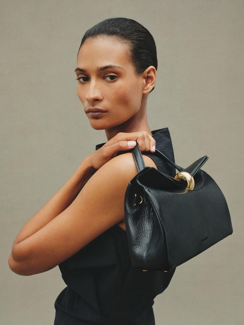 Elegant woman poses in a studio with a stylish black leather handbag, showcasing its gold accents and modern design against a neutral backdrop.