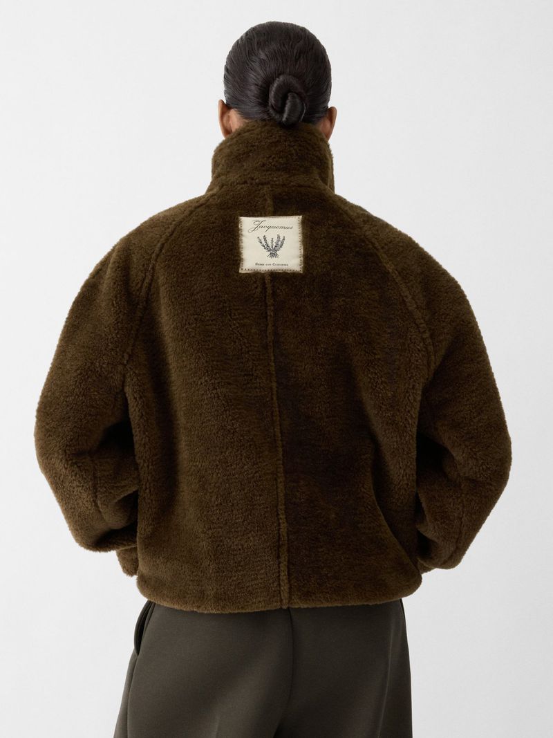 Back view of an adult woman with a bun, wearing an olive-brown sherpa jacket with a logo patch, and matching trousers on a white backdrop.