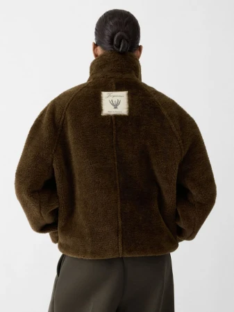 Back view of an adult woman with a bun, wearing an olive-brown sherpa jacket with a logo patch, and matching trousers on a white backdrop.