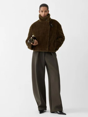 Model Mar G wearing a brown fleece jacket and wide-leg trousers holding the Jacquemus Le Valérie bag against a white backdrop.
