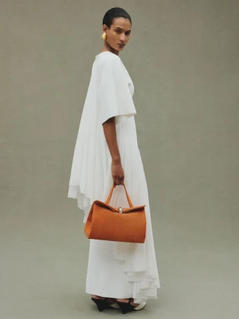 Model Mar G wearing a draped white dress and holding an orange suede Jacquemus Le Valerie handbag against a neutral studio backdrop.