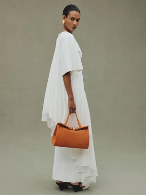 Model Mar G wearing a draped white dress and holding an orange suede Jacquemus Le Valerie handbag against a neutral studio backdrop.