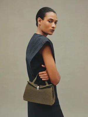 Model Mar G wearing a dark sleeveless dress and holding an olive green Jacquemus Le Valérie handbag against a neutral grey backdrop.