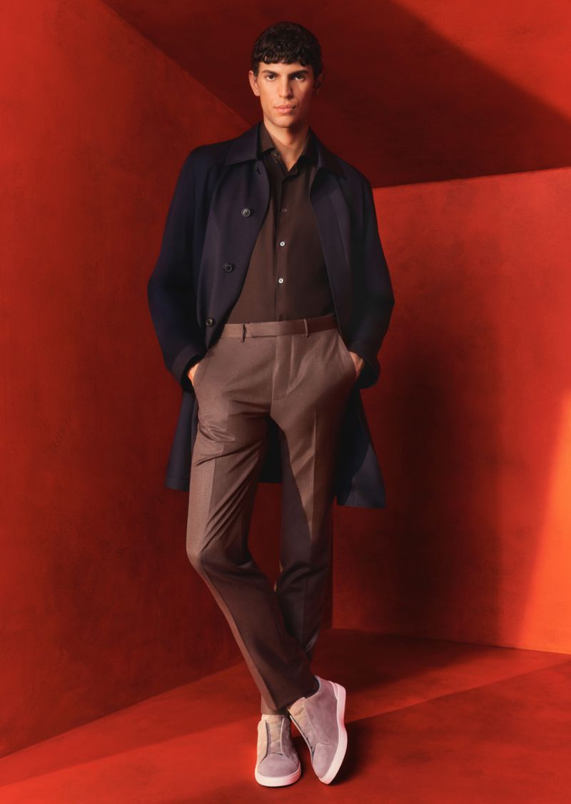 Fashionable man in a brown collared shirt and trousers, layered with a dark blue coat, standing against a red background in a studio.