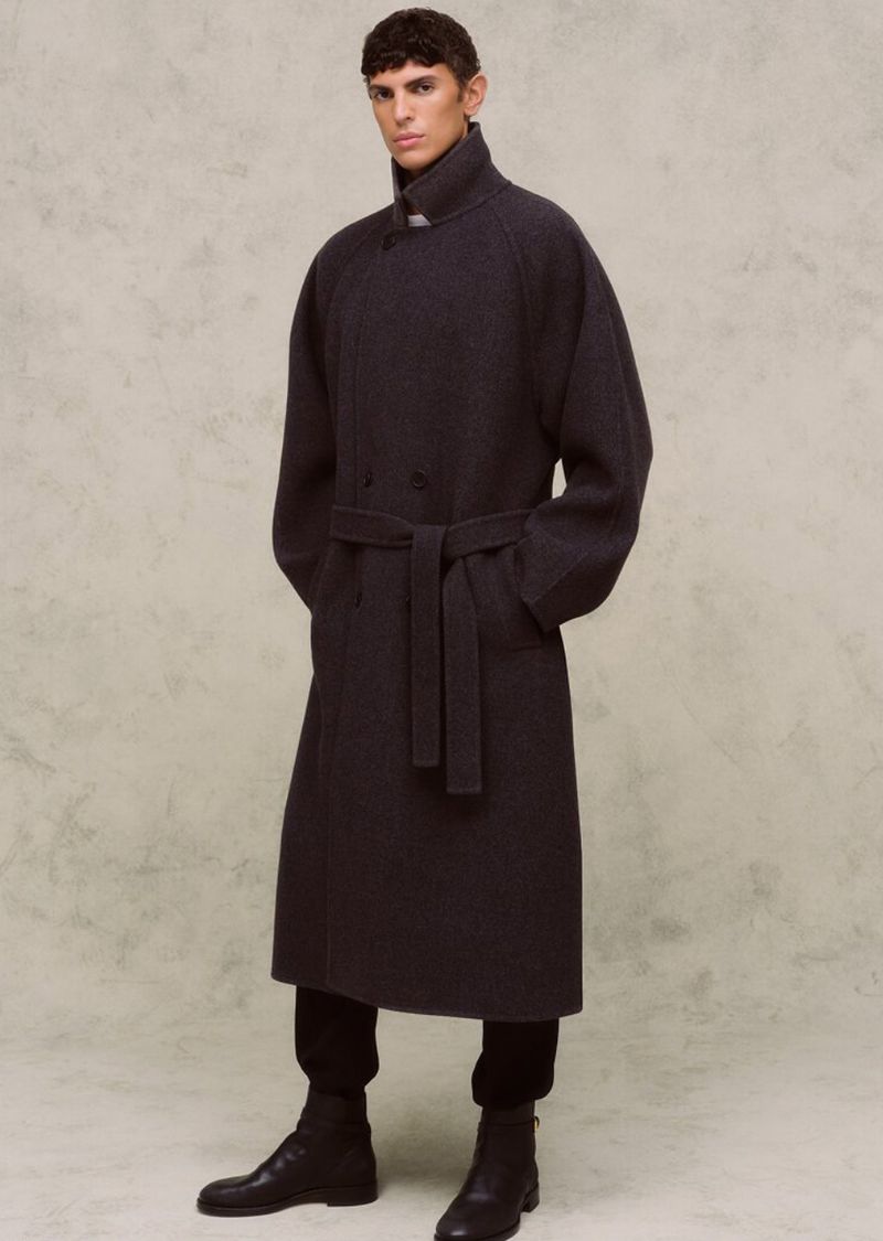 A single adult male standing indoors wearing a long, belted wool coat, black pants, and black leather ankle boots against a neutral backdrop.