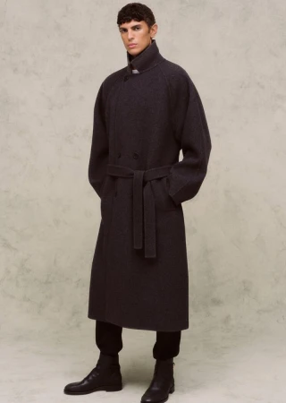 A single adult male standing indoors wearing a long, belted wool coat, black pants, and black leather ankle boots against a neutral backdrop.