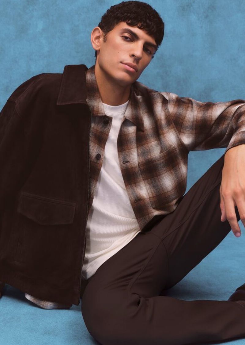 Adult man in a brown suede jacket with a plaid shirt and dark pants sitting in a photo studio with a blue textured background.