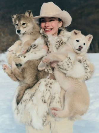 Woman in hat and fur coat smiling, holding three wolf pups in snow, medium shot, sunny day.