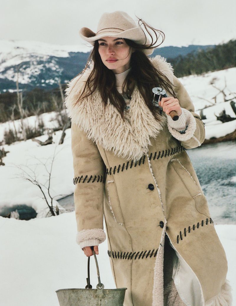 A woman in a cowboy hat and shearling coat stands in a snowy landscape, holding a metal bucket and fishing reel under an overcast sky.