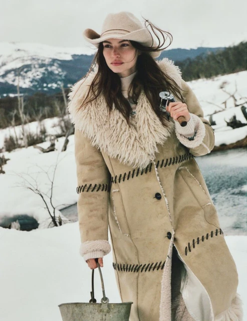 A woman in a cowboy hat and shearling coat stands in a snowy landscape, holding a metal bucket and fishing reel under an overcast sky.