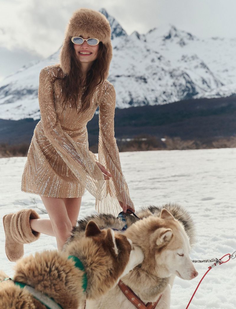A smiling woman in a sequin dress and fur hat poses with sled dogs against a snowy mountain backdrop in an outdoor winter scene.