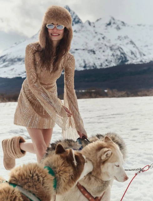 A smiling woman in a sequin dress and fur hat poses with sled dogs against a snowy mountain backdrop in an outdoor winter scene.
