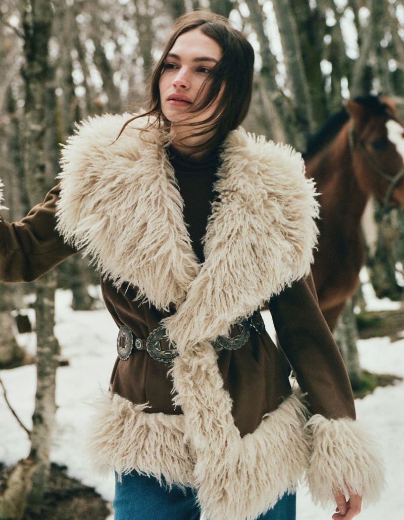 A woman in a furry coat with a horse in a snowy forest setting. The woman is looking to the side, in an outdoor setting.