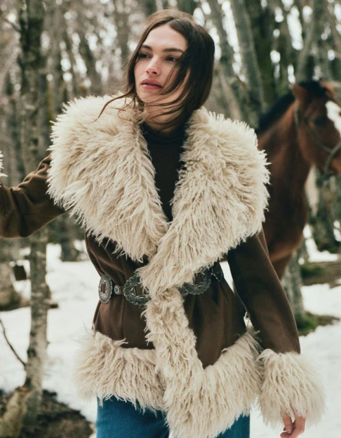 A woman in a furry coat with a horse in a snowy forest setting. The woman is looking to the side, in an outdoor setting.